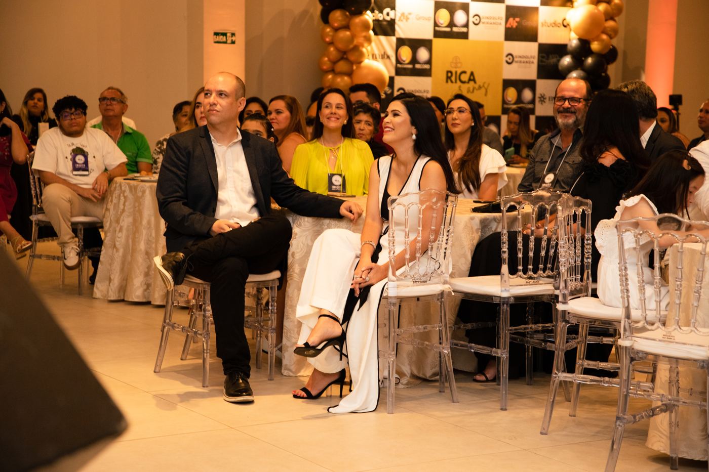 Plateia atenta em cadeiras de acrílico, com balões dourados ao fundo.
