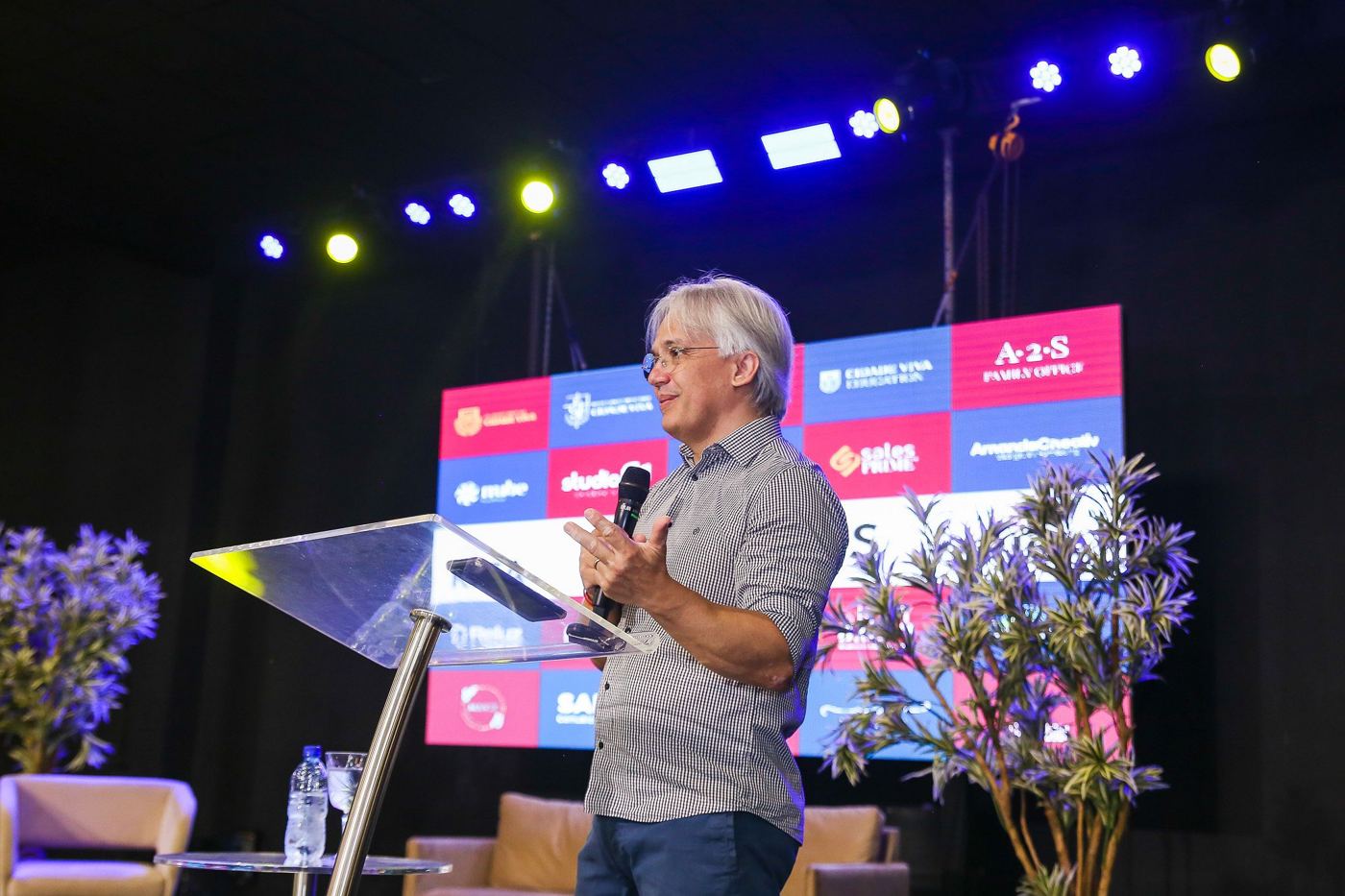Palestrante no palco, com flores e o painel das empresas parceiras ao fundo.