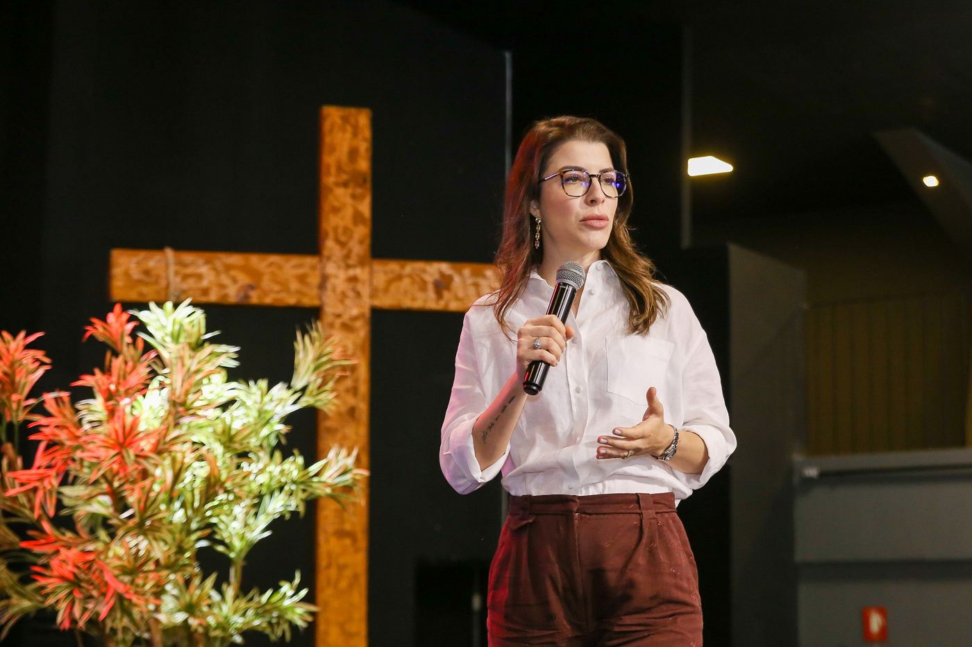 Palestrante ao microfone no palco, com uma cruz de madeira e flores ao fundo.